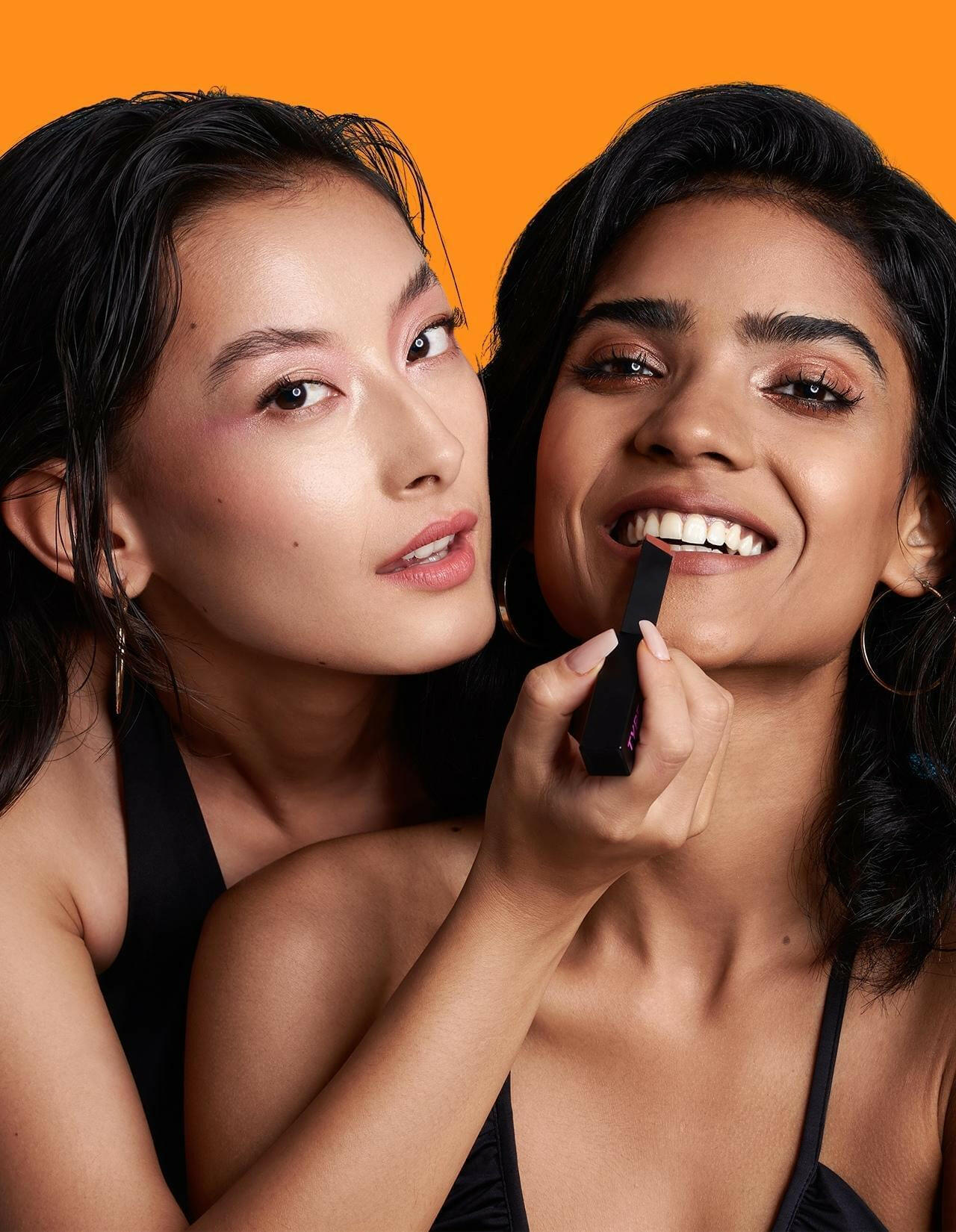 Two women applying SOAK IT LIPSTICK against a vibrant orange background.